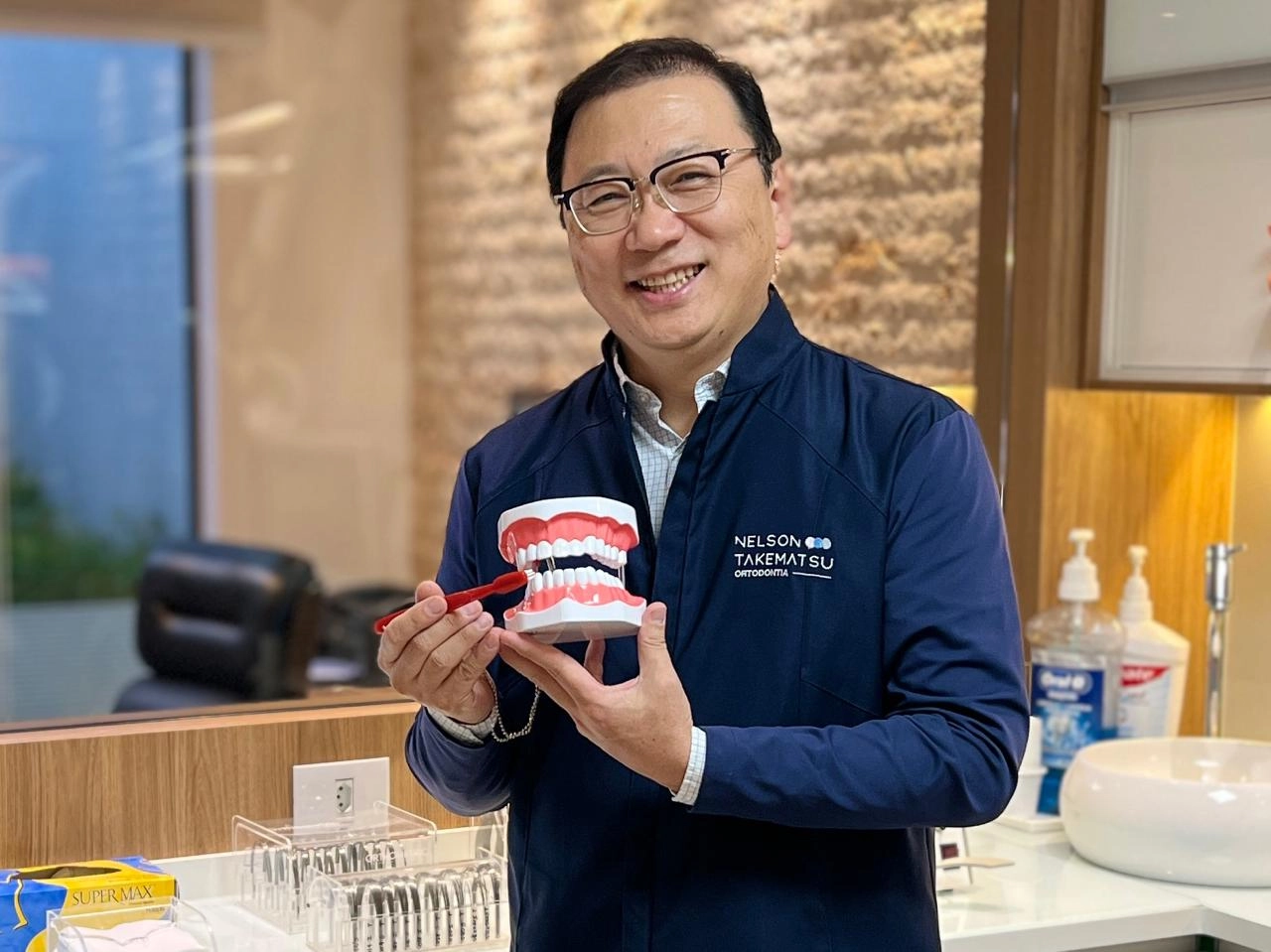 Dr. Nelson Takematsu apresentando macro modelo dentário com escova — ortodontia infantil e preventiva em Mogi das Cruzes